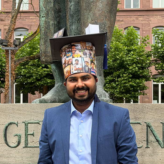 Smiling man in a blue suit and a graduation cap decorated with photos, standing before a statue.