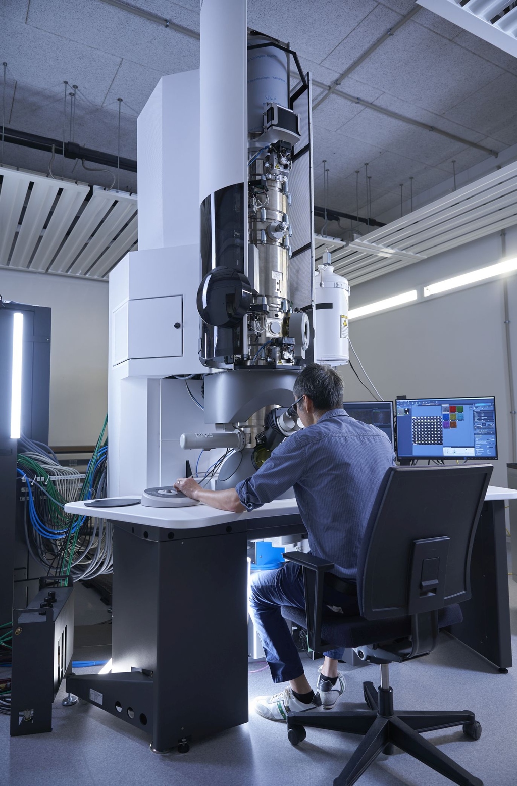 Scientist at a high-tech electron microscope, viewing screens, in a lab.